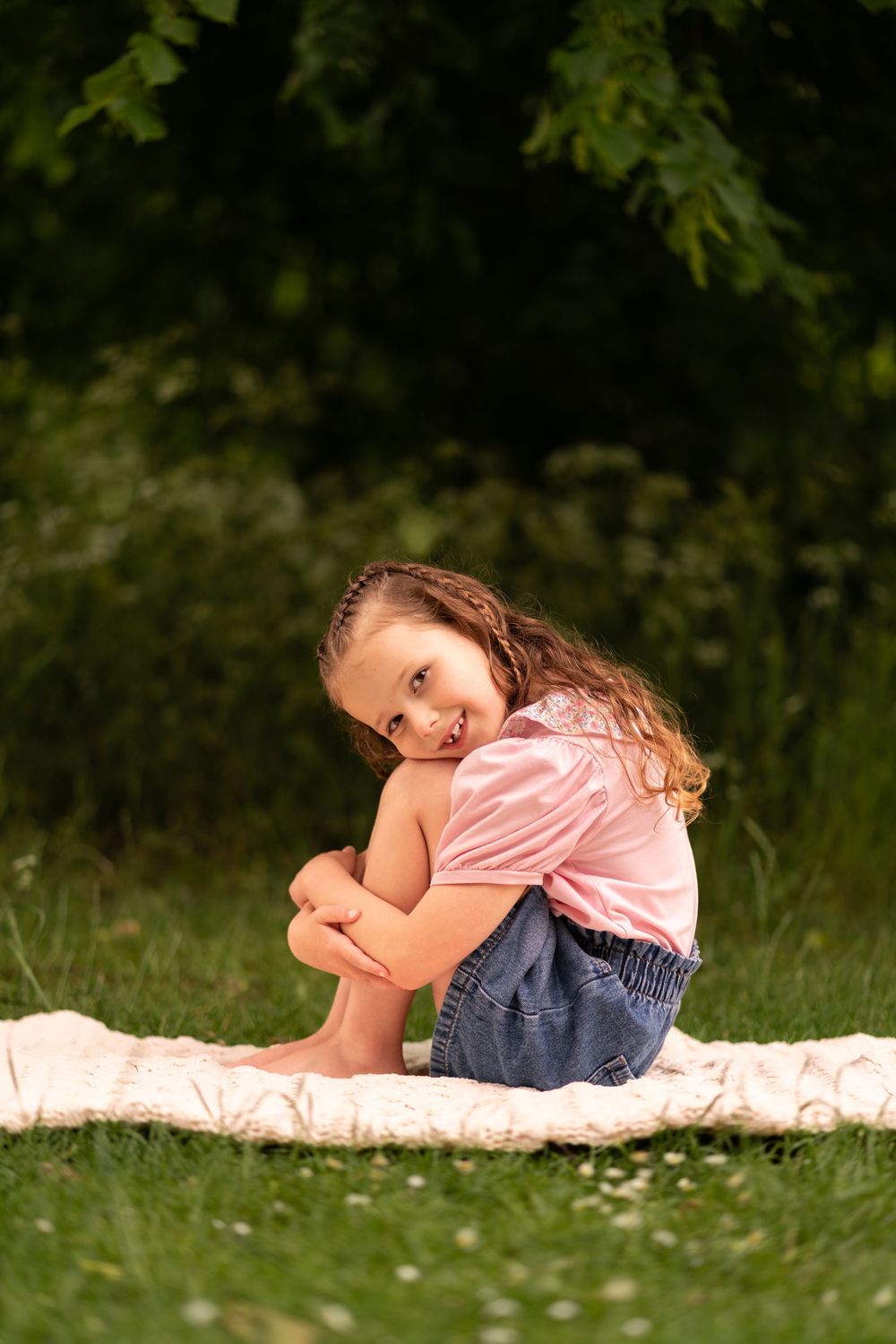 A young girl sits on a blanket outdoors, hugging her knees and smiling during a childhood photoshoot in Lincolnshire.