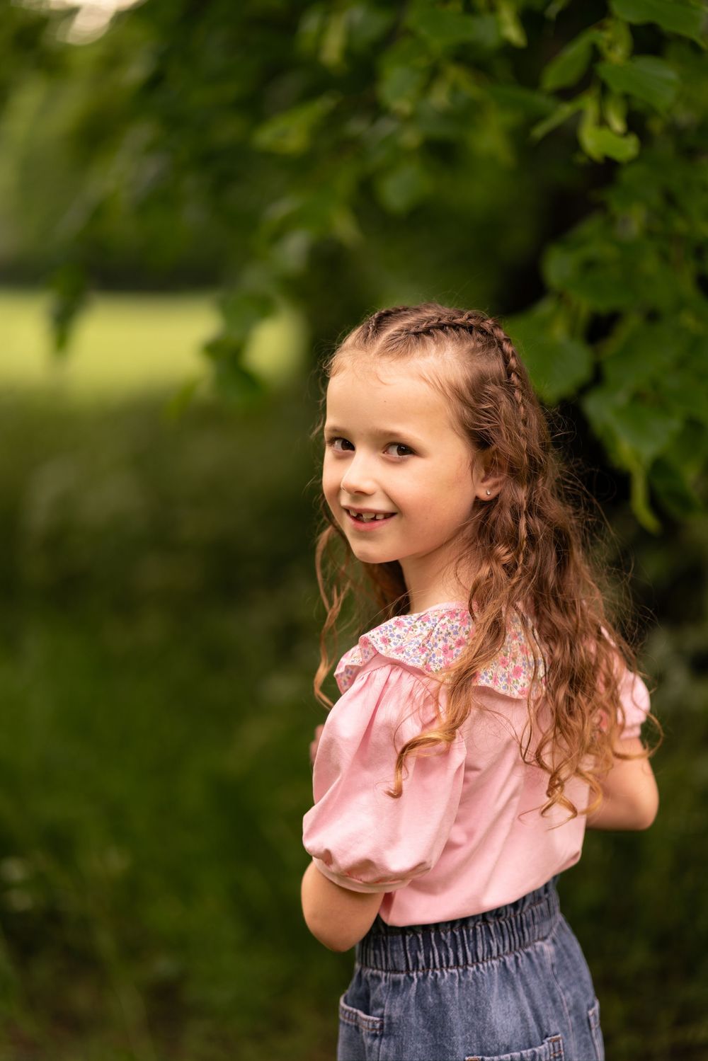 A smiling young girl with curly hair in a pink blouse and denim skirt poses outdoors during a childhood photography session in Lincolnshire.