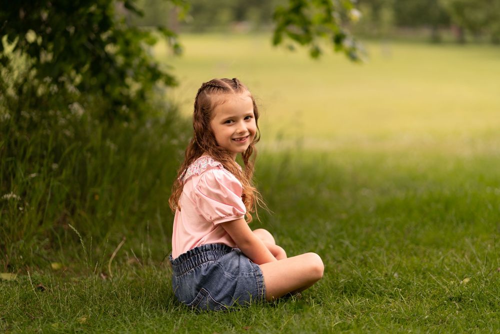A smiling girl sits cross-legged on green grass in a park, wearing a pink top and denim shorts during a childhood photoshoot in Lincolnshire