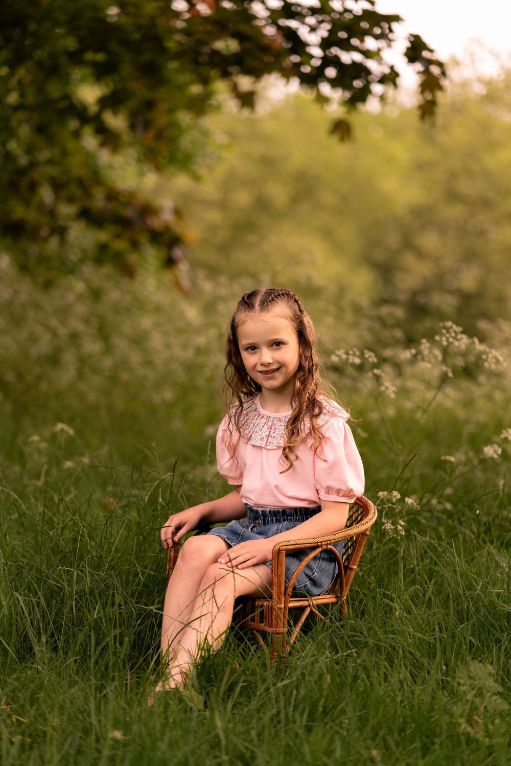 Young girl smiling while seated in wicker chair outdoors in lush green meadow during childhood photoshoot in Lincolnshire.