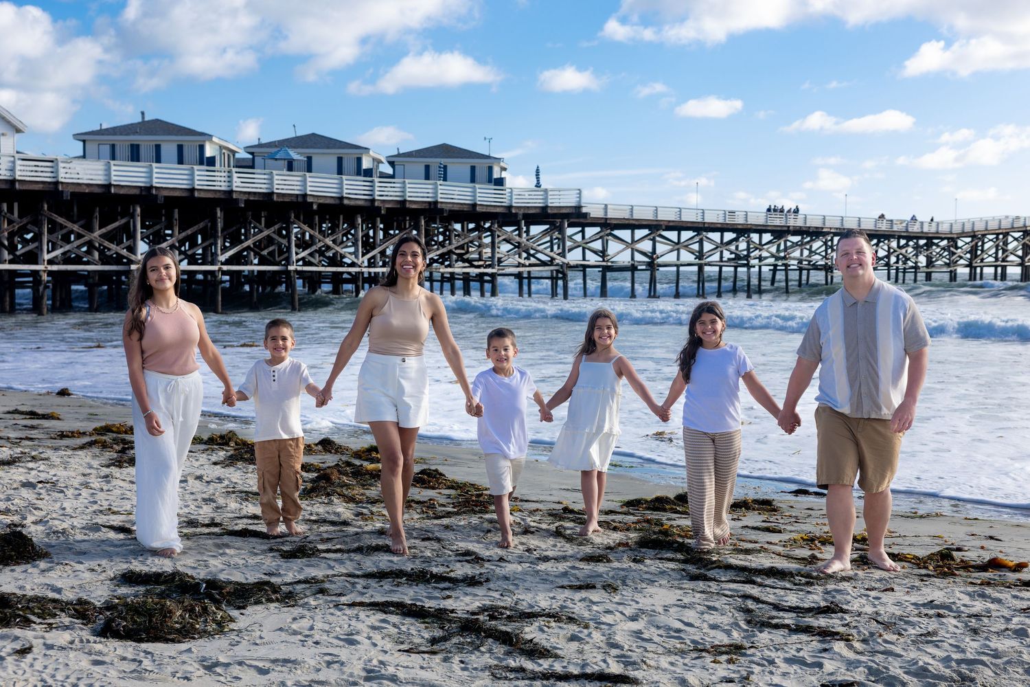 San Diego family portrait photography of a family holding hands on a beach in front of Crystal Pier with ocean waves.