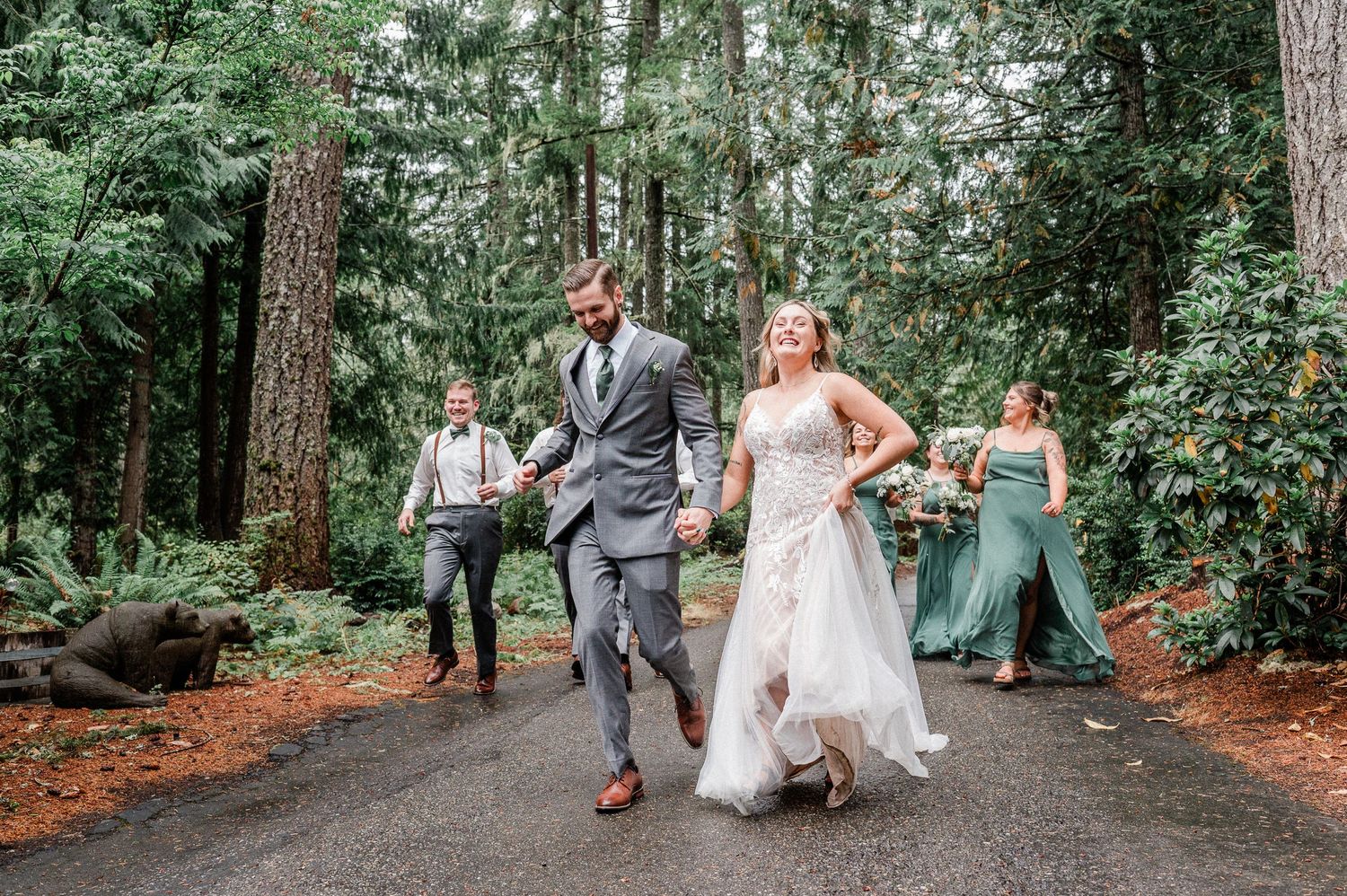 Bride and groom run hand-in-hand on forest path with wedding party in lush green woodland setting.