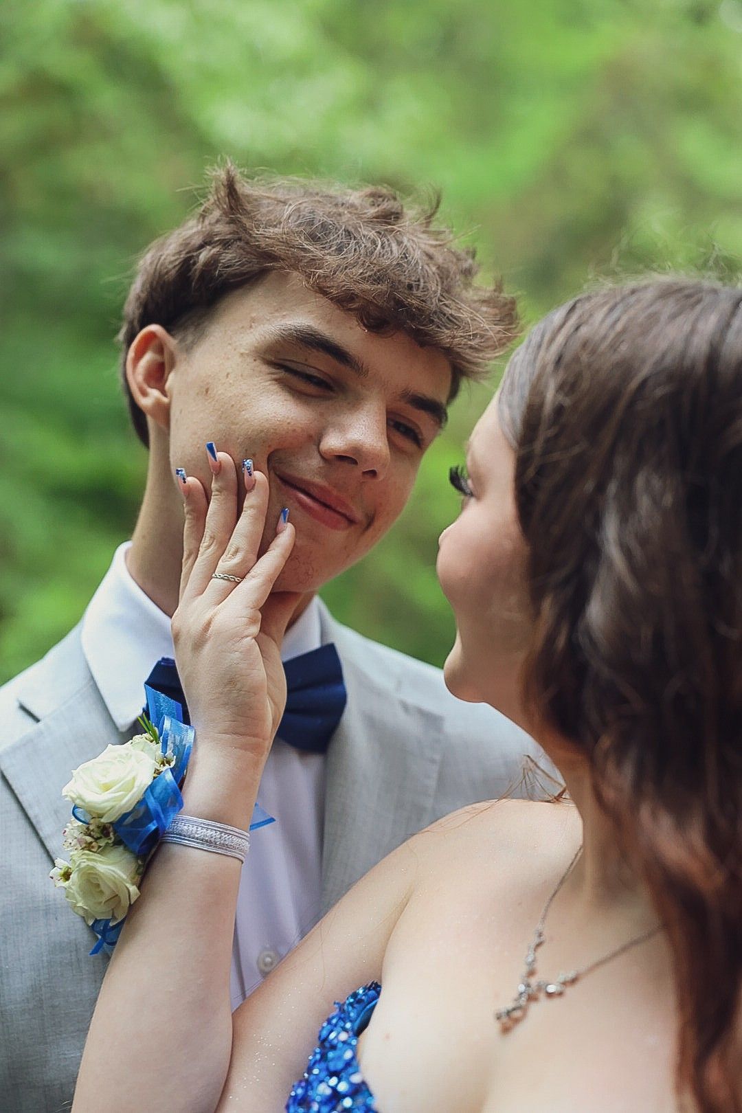 Prom couple posing by stone wall in forest, woman in royal blue gown, man in gray suit.