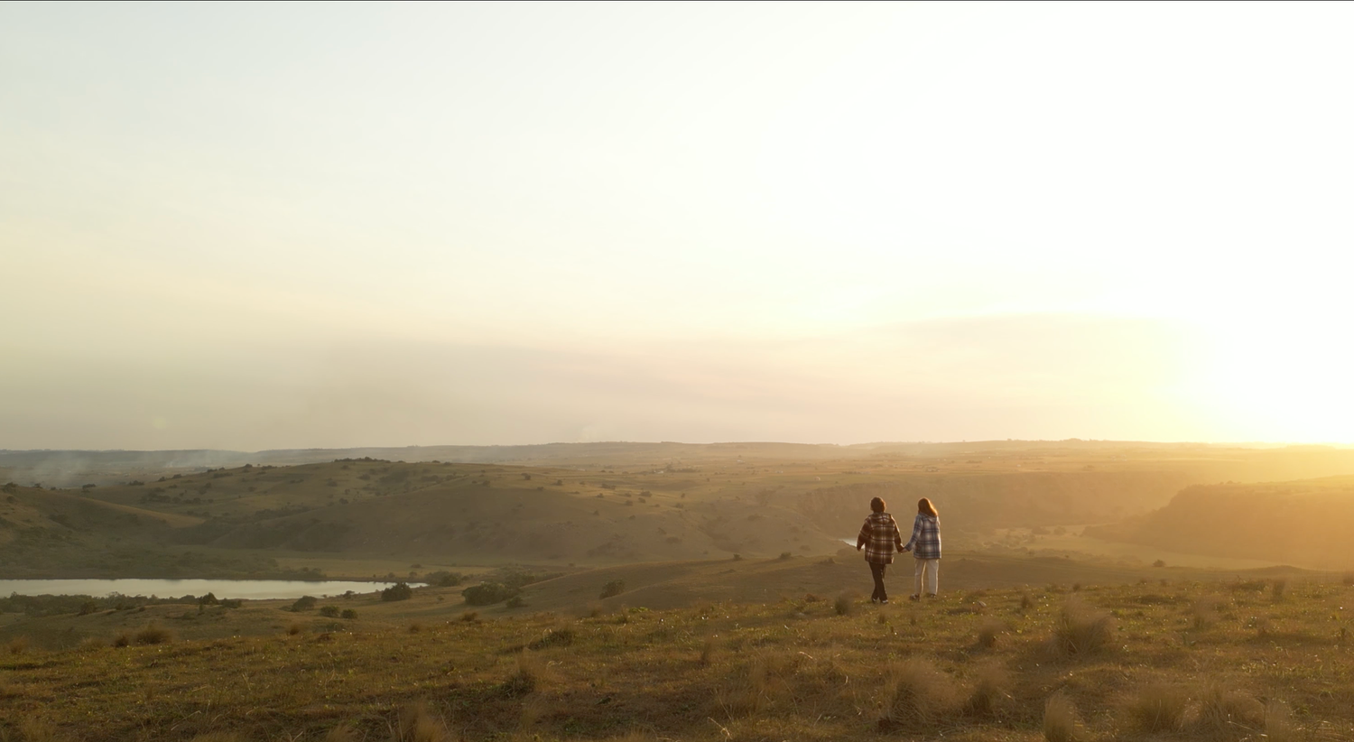 Two people walking on a scenic hilltop landscape at golden sunset near a river.