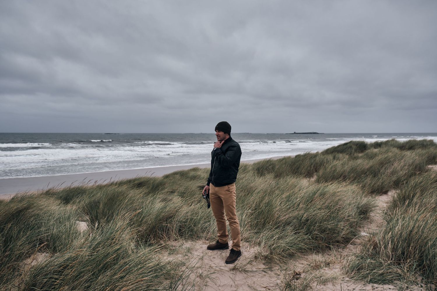 Man standing on sandy beach dunes with tall grass, overlooking a moody cloudy ocean shoreline.