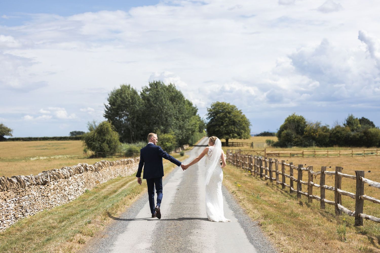 Bride and groom walk hand in hand down a rural country lane flanked by stone walls and wooden fences.