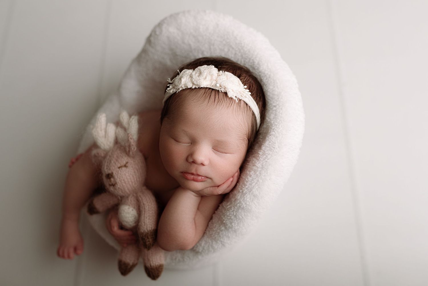 Sleeping newborn baby in white fluffy wrap holding a knit bunny toy, wearing a floral headband.