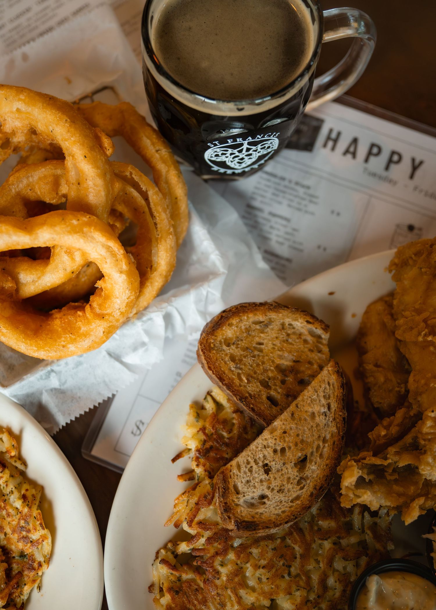 Overhead view of onion rings, hash browns, toast, fried chicken, and dark beer mug on a diner table with happy hour menu.