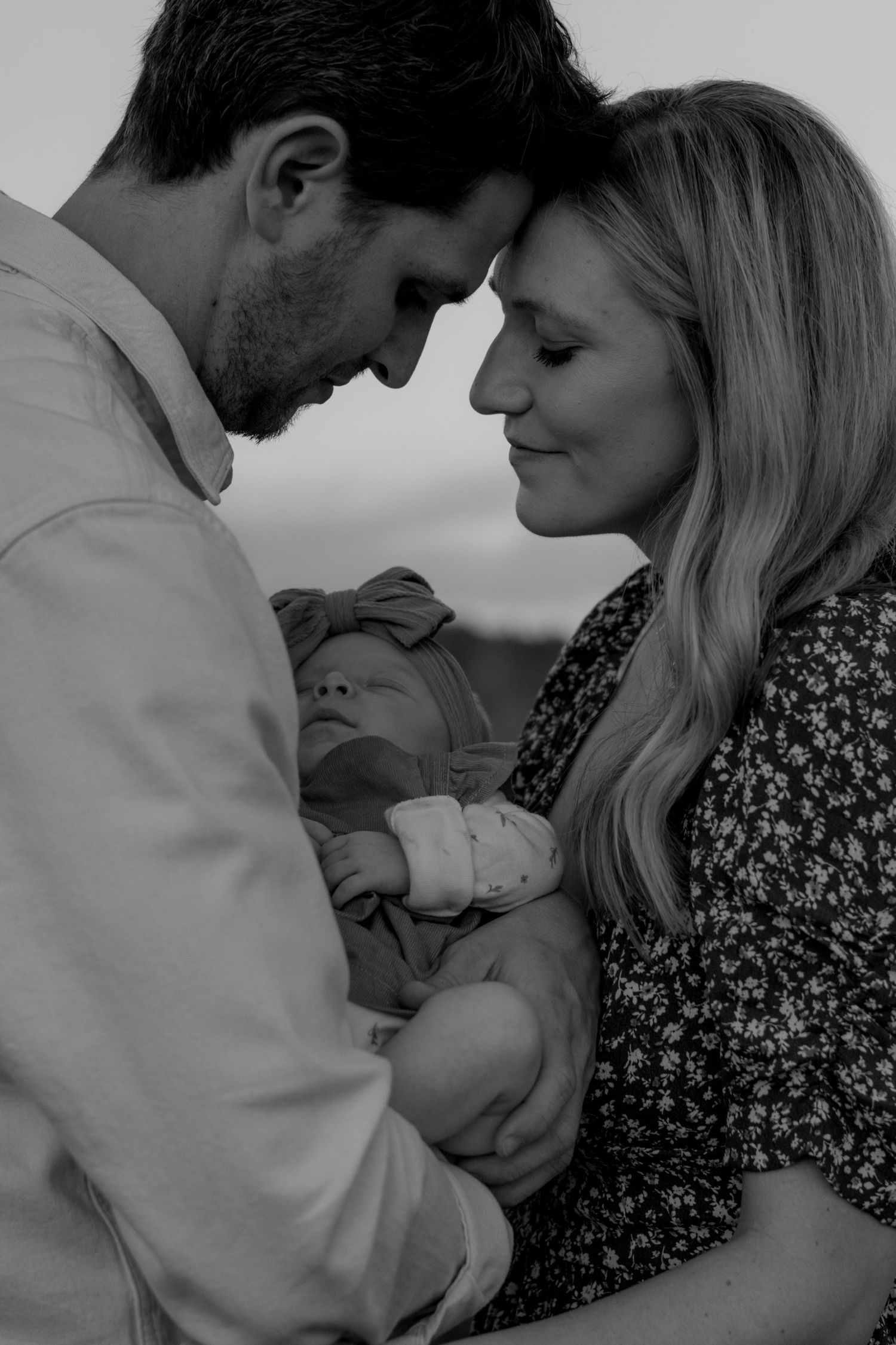 Tender black and white portrait of new parents touching foreheads while cradling their newborn baby.