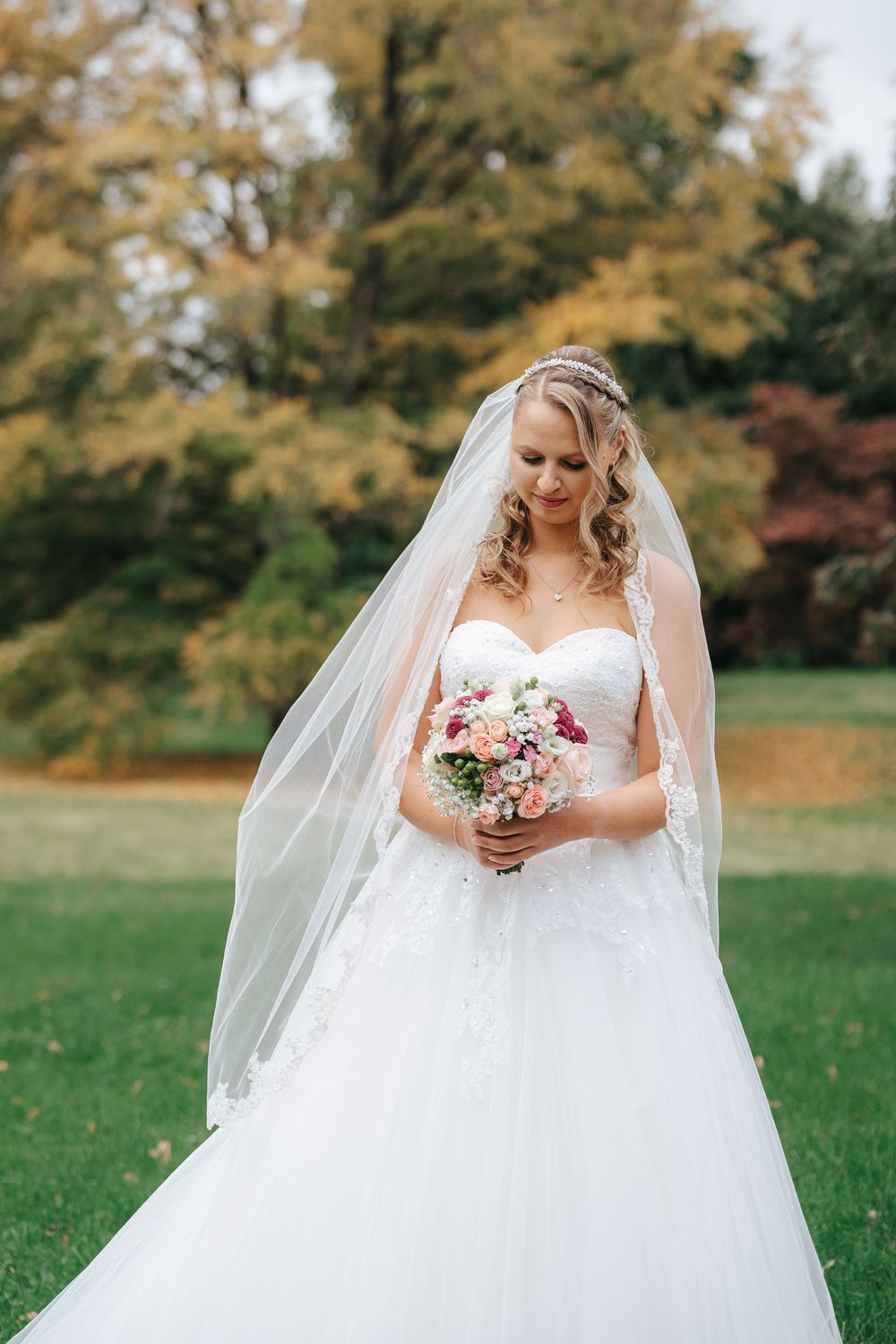 Bride in white lace ballgown and veil holds floral bouquet outdoors with autumn trees in background.