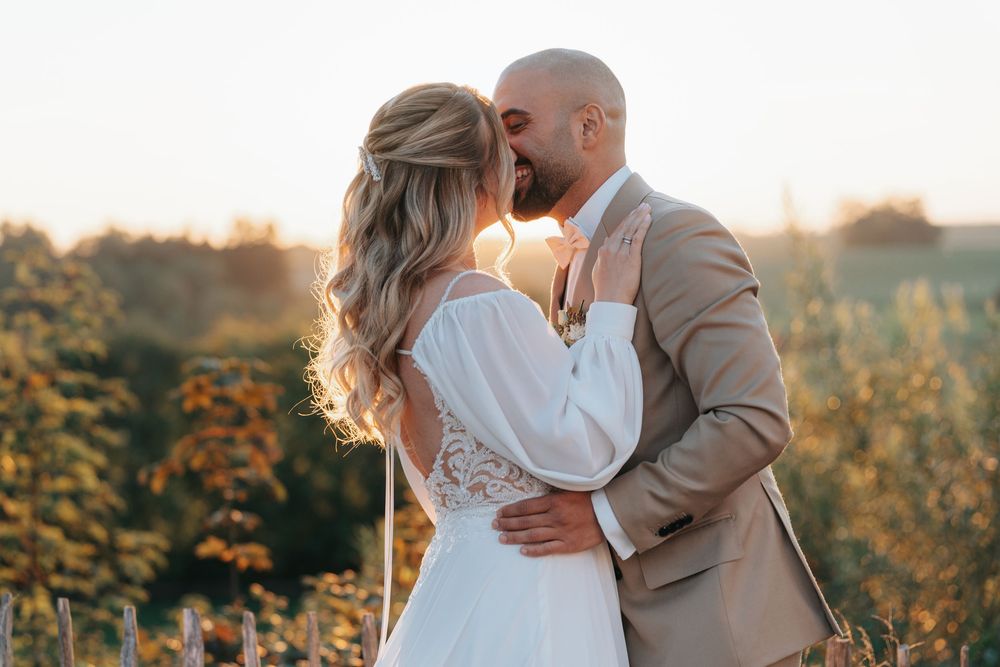 Bride and groom share a romantic kiss at golden hour in a sunlit field, bride in white lace gown, groom in tan suit.