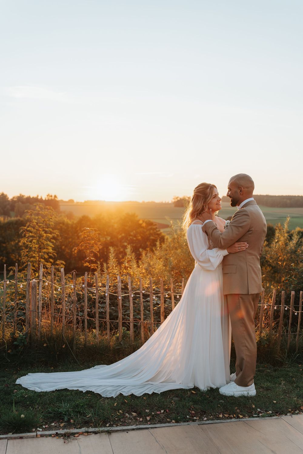 Bride and groom embrace at golden sunset overlooking lush countryside, bride in flowing white gown, groom in tan suit.