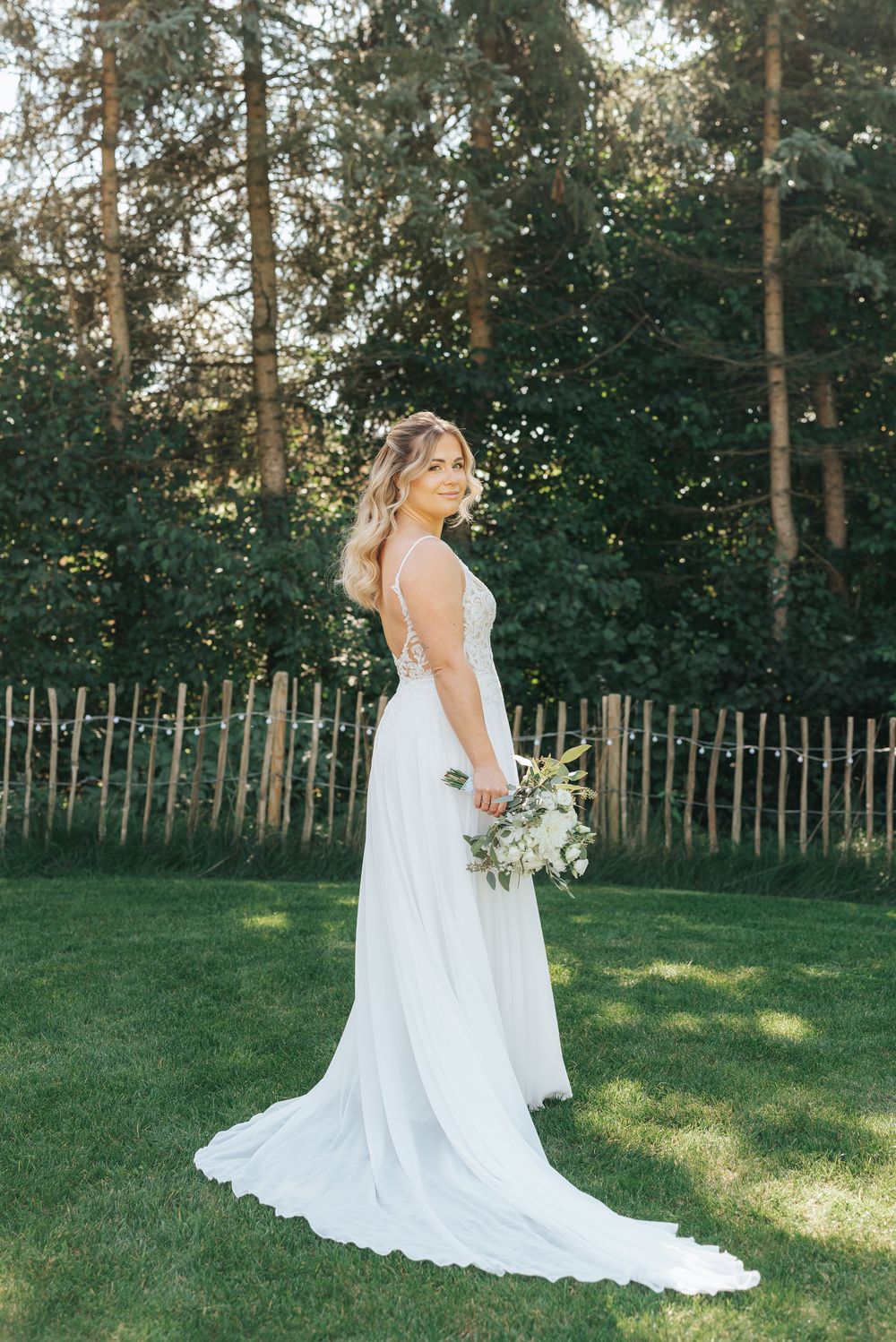 Bride in a flowing white wedding gown holding a bouquet, standing on green grass with a wooden fence and trees behind her.