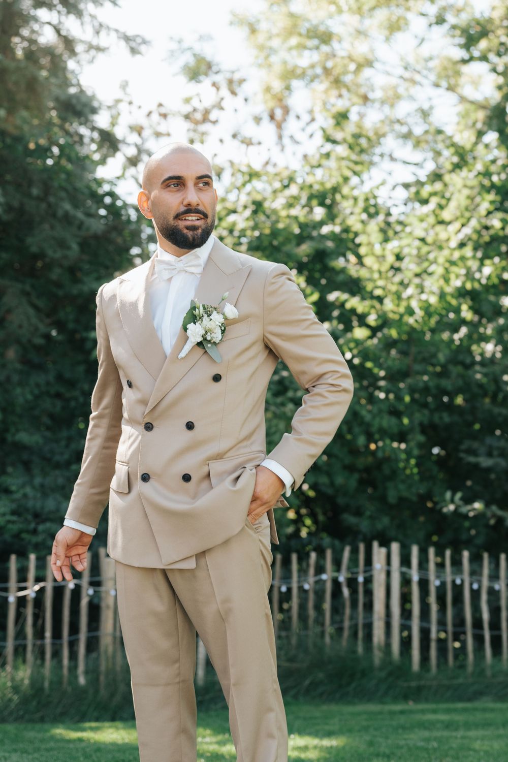 Bald groom in beige double-breasted suit with white boutonniere stands confidently outdoors at garden wedding.