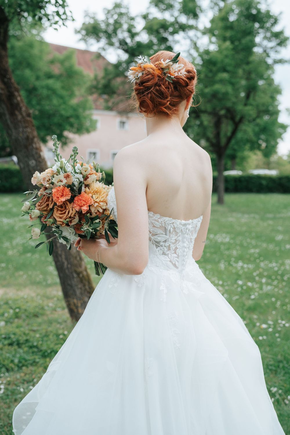 Bride in white strapless gown holding orange floral bouquet, red hair updo, standing in green garden.