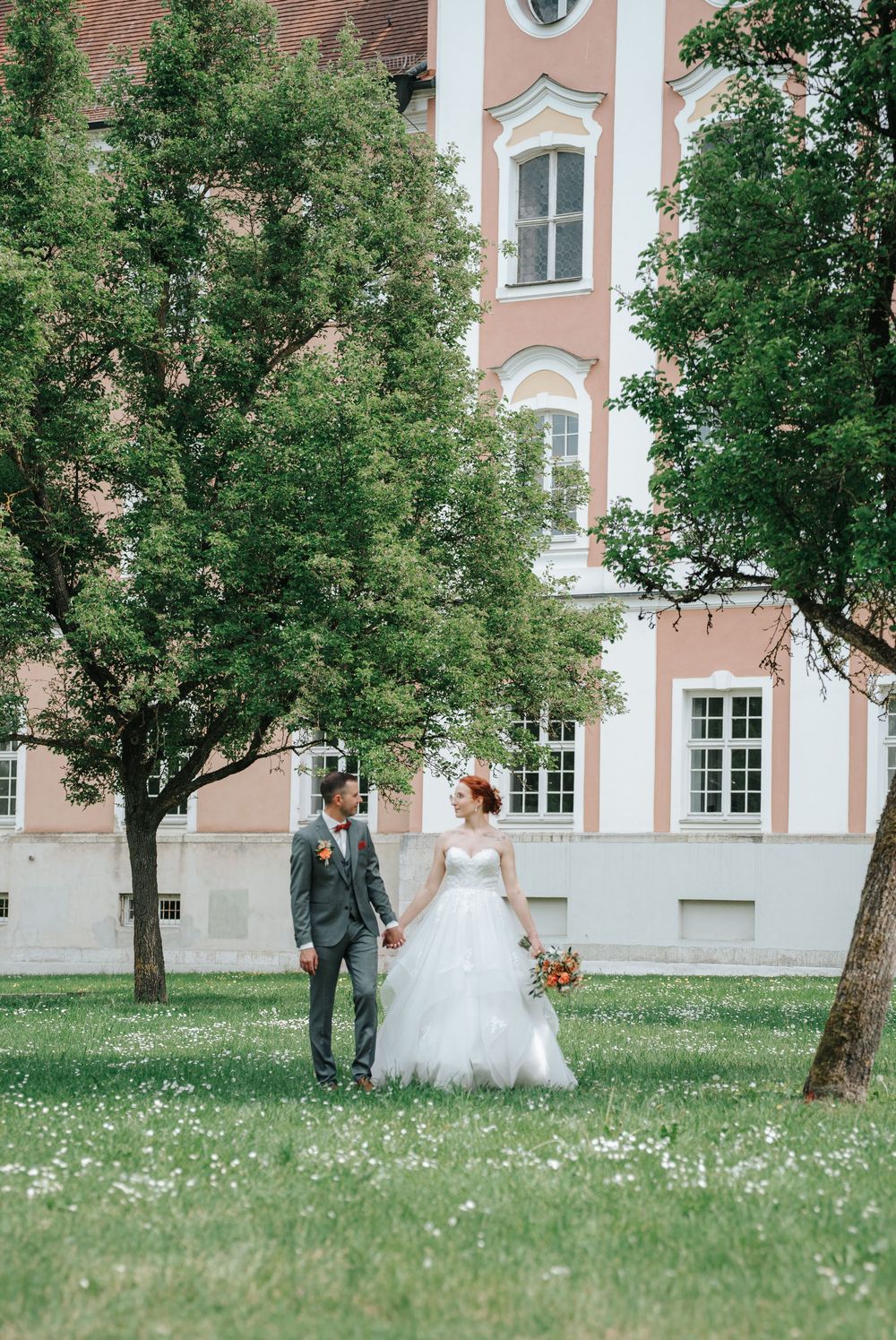 Bride and groom holding hands walking on green grass lawn beside trees with elegant pink historic building backdrop.