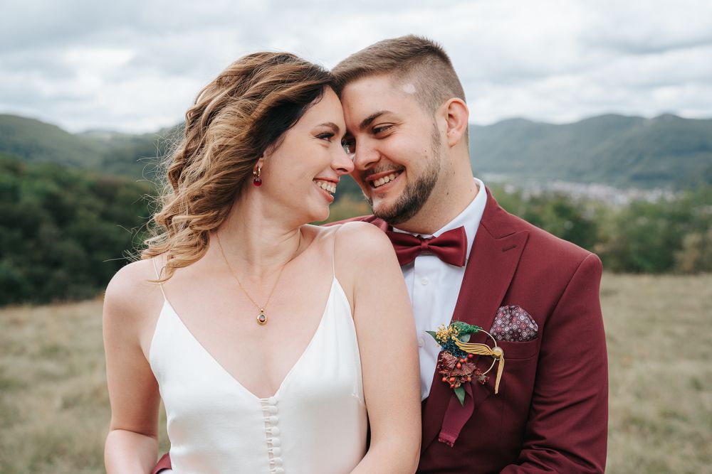 Bride and groom smiling closely together outdoors, wearing white dress and burgundy suit with bow tie and boutonniere.