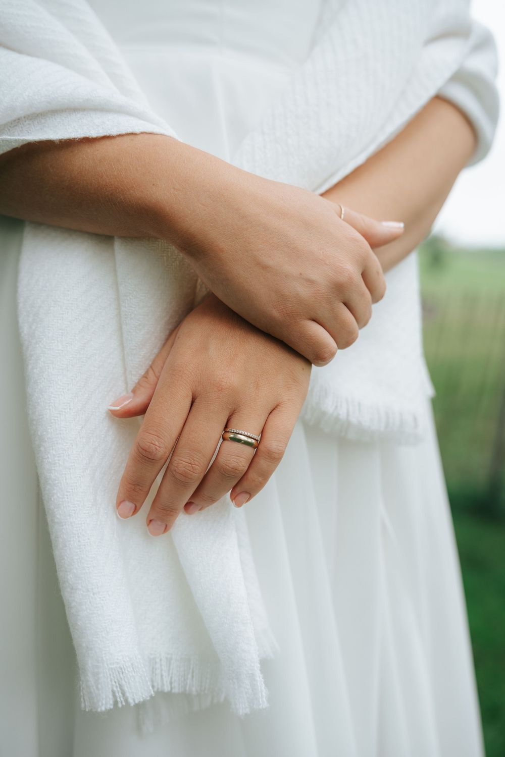 Close-up of bride's hands crossed over white dress, wearing a gold wedding band with soft pink manicure.