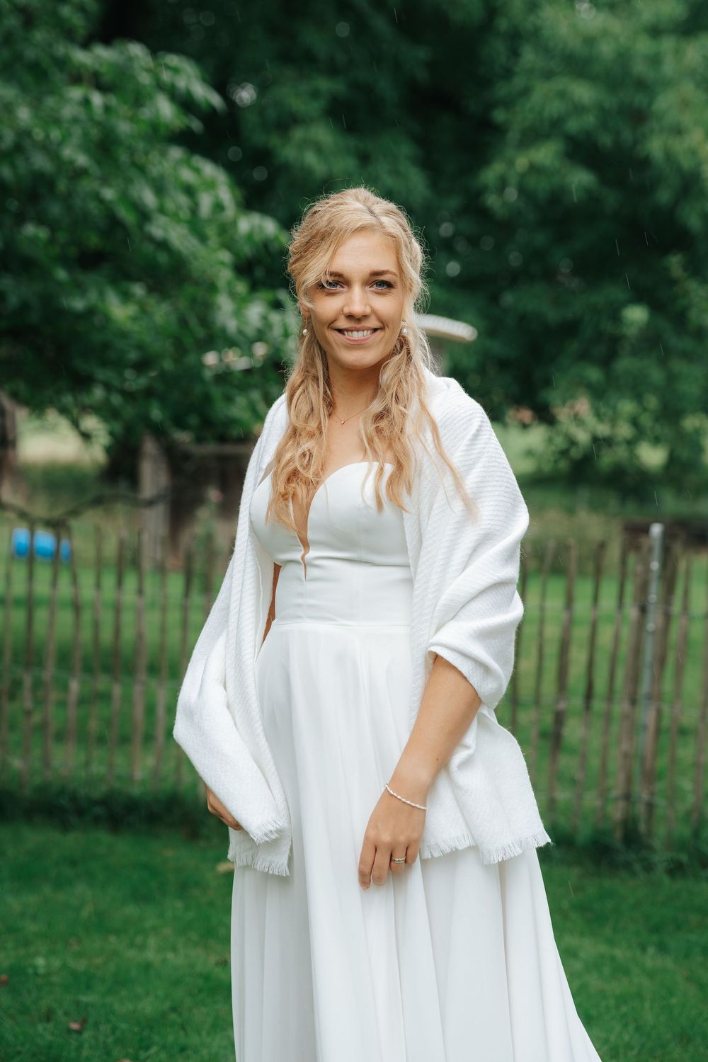 Smiling blonde bride in white dress and shawl posing outdoors in lush green garden setting.