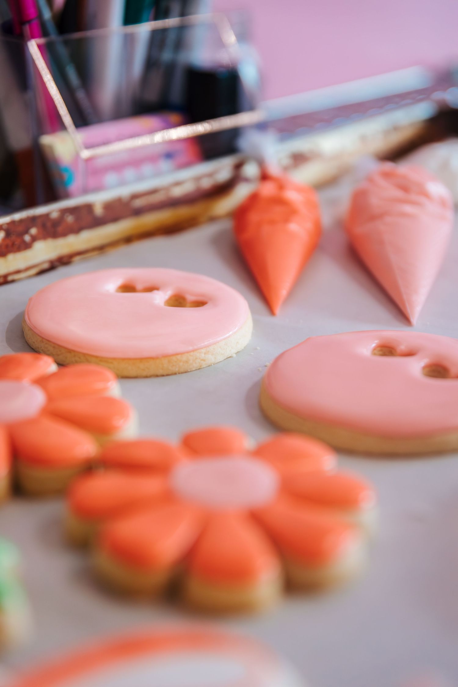 Pink and orange iced sugar cookies shaped as buttons and flowers on parchment paper with decorating bags nearby. Bakery branding photoshoot