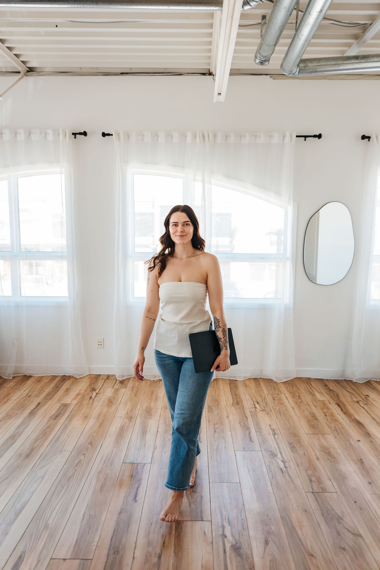 Woman in strapless top and jeans holding a laptop, standing barefoot on hardwood floors in a bright studio space. Business pictures