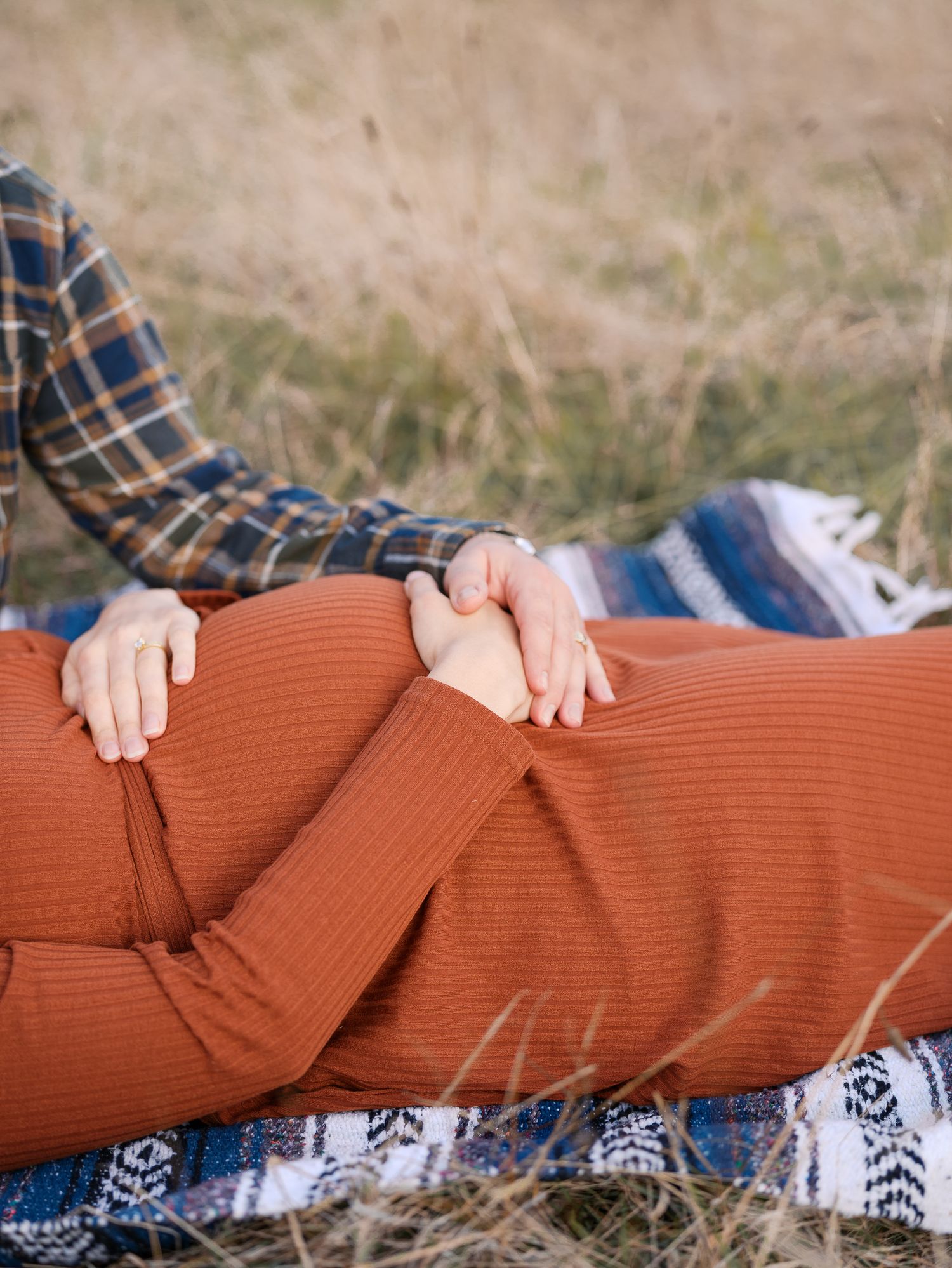 Pregnant couple lying on a blanket in a field, hands resting on baby bump, wearing rust and plaid outfits.