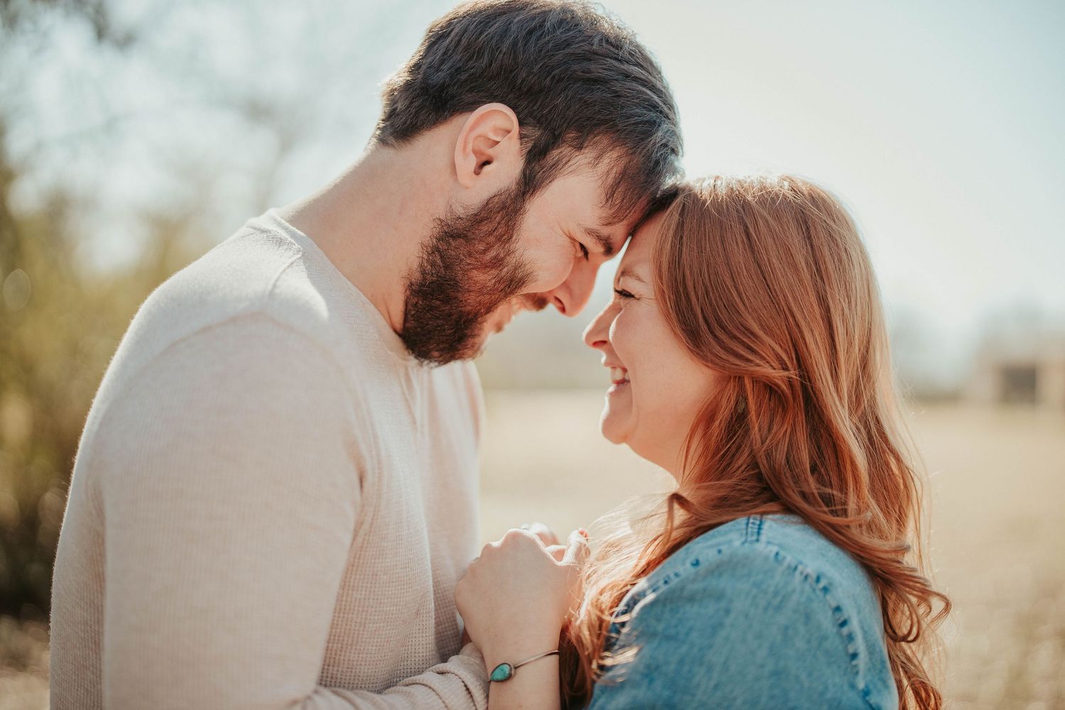 Romantic couple touching foreheads and holding hands outdoors in a sunny field, sharing an intimate moment.