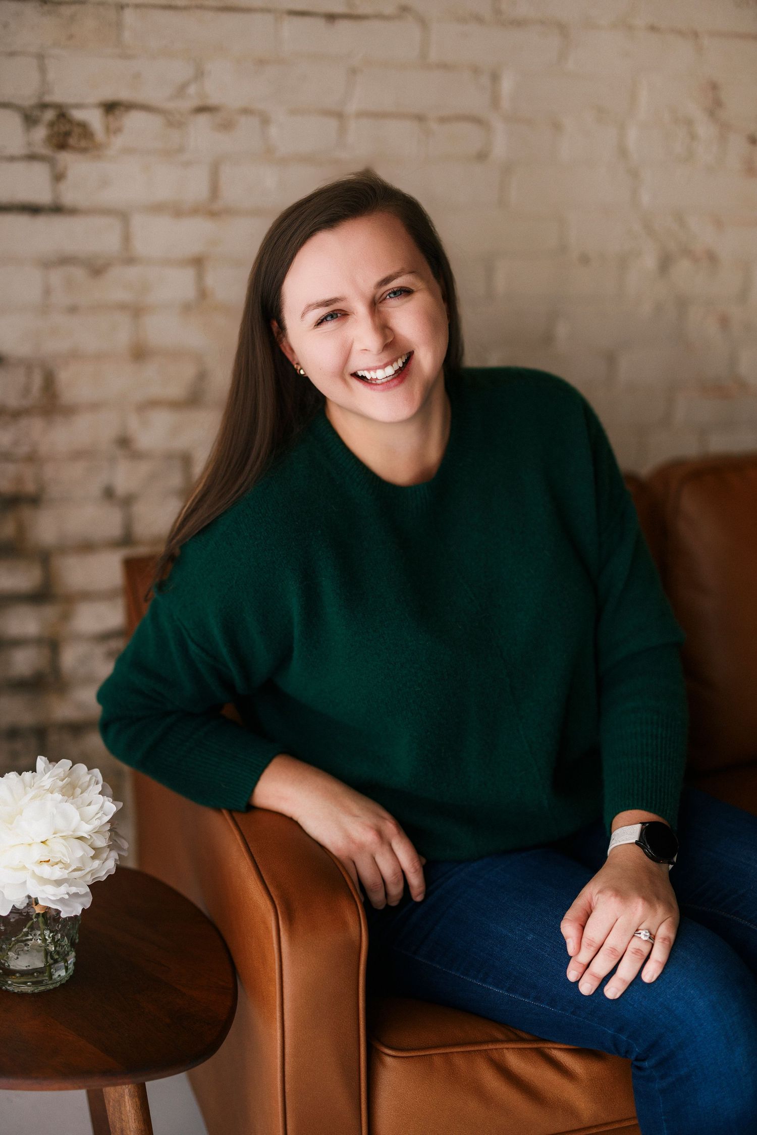 Smiling woman in green sweater and jeans sitting in brown leather chair against brick wall.