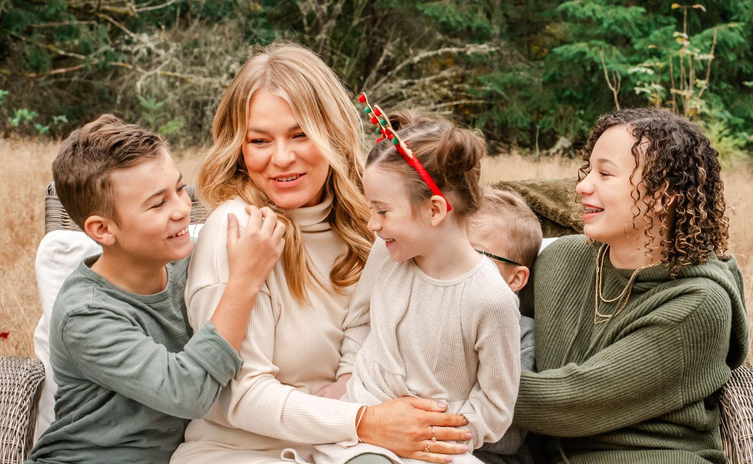 Smiling blonde woman surrounded by four happy children laughing outdoors in a natural wooded setting.