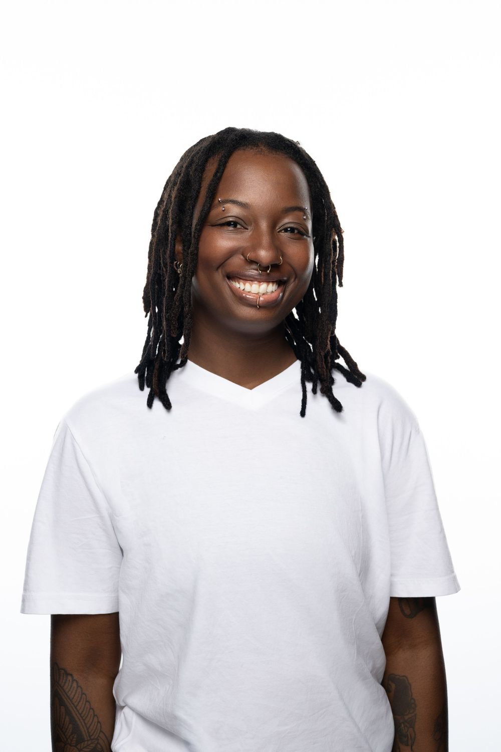 Smiling person with dreadlocks wearing a white v-neck t-shirt against a white background.