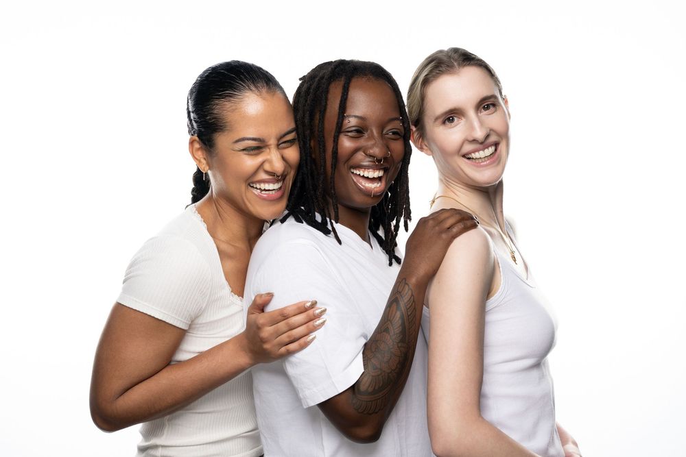Three diverse women in white tops smiling and laughing together against a white background.