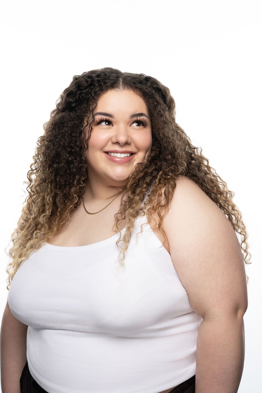 Smiling young woman with long curly hair wearing a white tank top posing against a white background.
