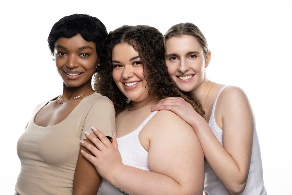 Three diverse women smiling together in casual tank tops against a white background, celebrating body positivity.