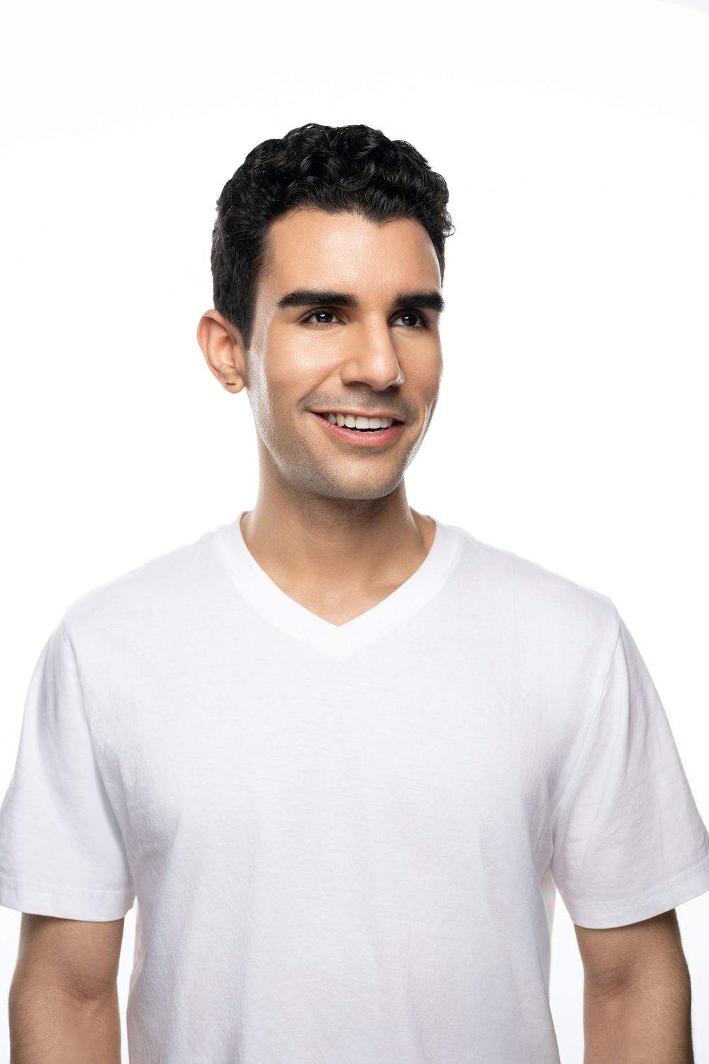 Smiling young man with dark curly hair wearing a white v-neck t-shirt against a white background.