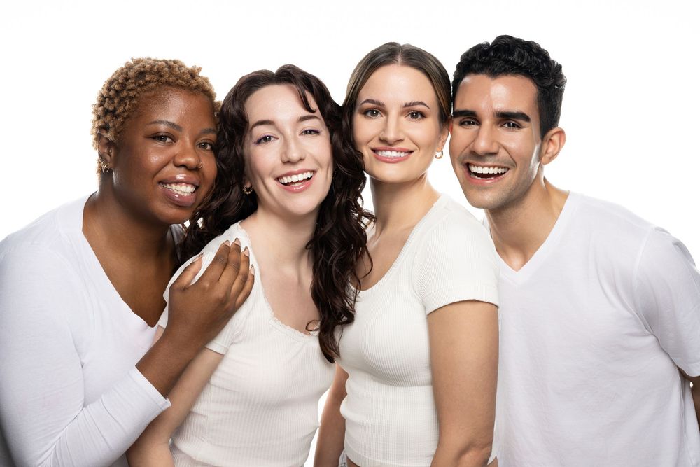 Four diverse smiling friends posing together in white tops against a white background.
