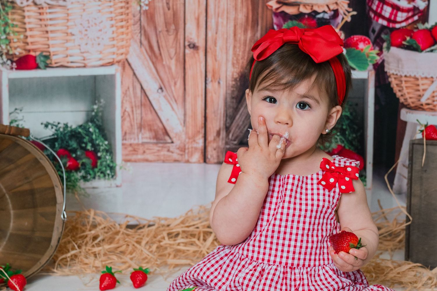 Cute toddler girl in red gingham dress eating strawberries in a rustic barn-themed photo setting.