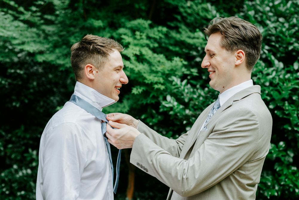 Groomsman adjusts groom's tie outdoors before wedding ceremony surrounded by lush green trees.