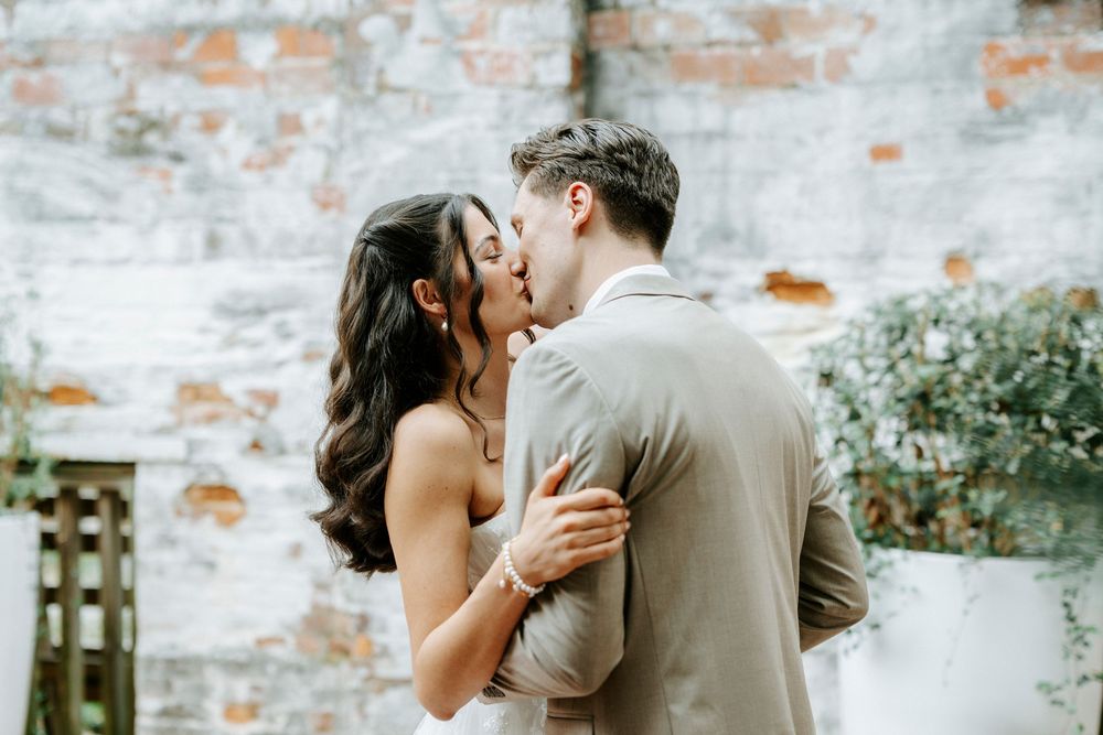A bride and groom share a romantic first kiss at their wedding ceremony in front of a rustic brick wall backdrop.