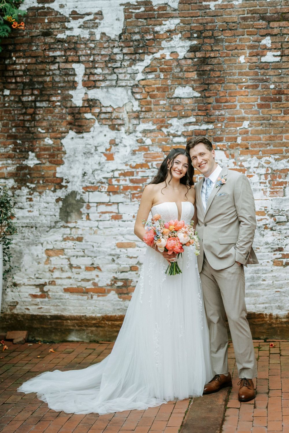 Bride and groom posing together in front of a rustic brick wall, bride holding a coral bouquet.