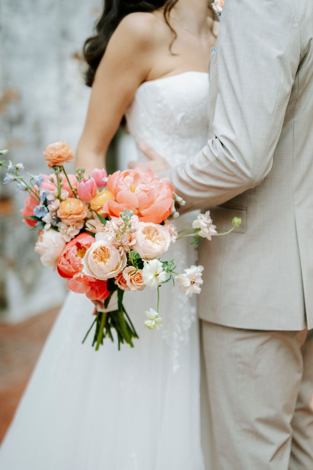 Bride and groom embracing while holding vibrant coral and peach wedding bouquet with garden roses.