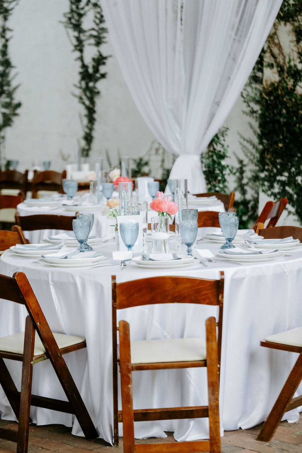 Elegant outdoor wedding reception table with white linen, blue glassware, pink floral centerpiece, and wooden chairs.