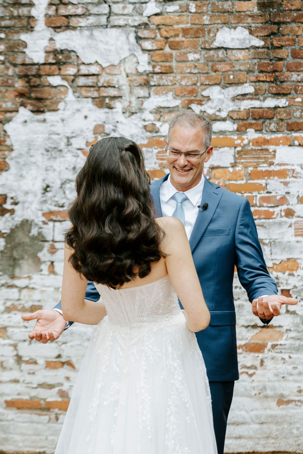 Emotional father reacts with joy during a first look with his daughter the bride against a rustic brick wall backdrop.