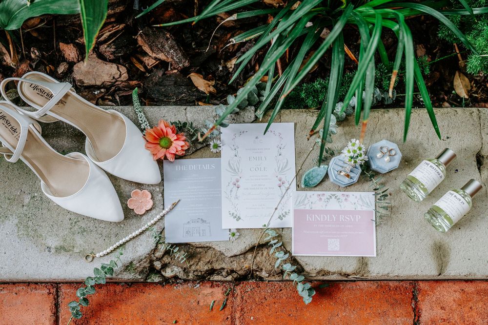 Elegant bridal flat lay featuring white heels, wedding invitations, perfume bottles, and floral accents on brick surface.