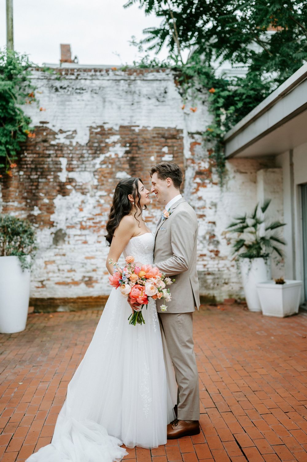 Bride and groom share a romantic moment in a rustic brick courtyard, bride holding a vibrant coral and pink bouquet.