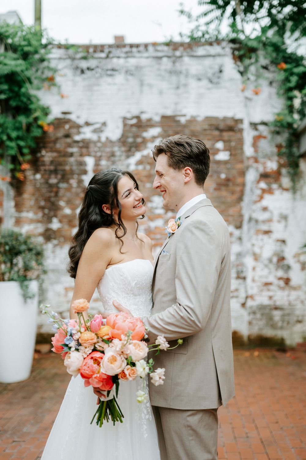 Bride and groom pose together smiling, bride holding vibrant coral bouquet against rustic brick wall.