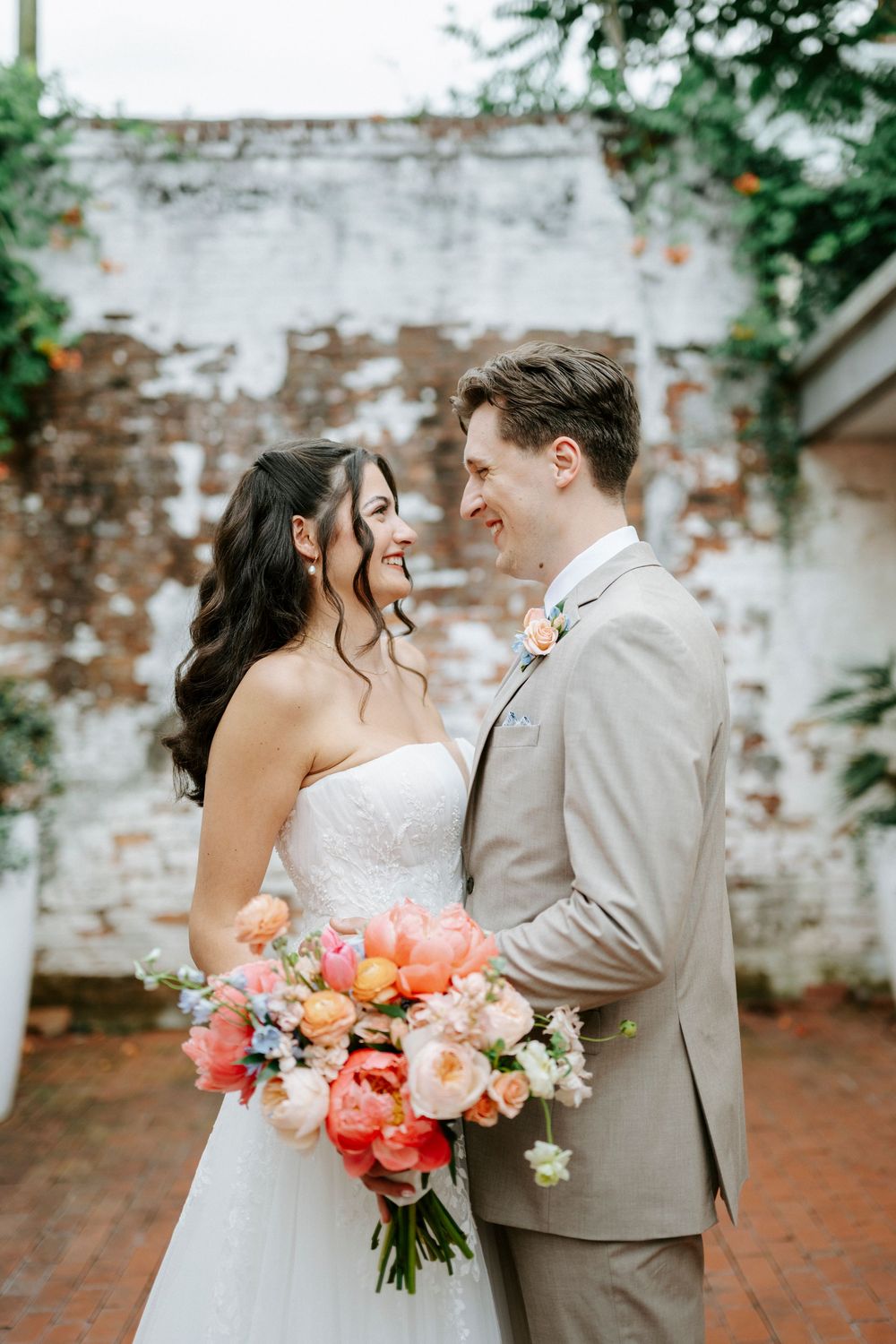 Smiling bride in strapless gown and groom in tan suit pose together holding colorful peony bouquet at outdoor wedding.