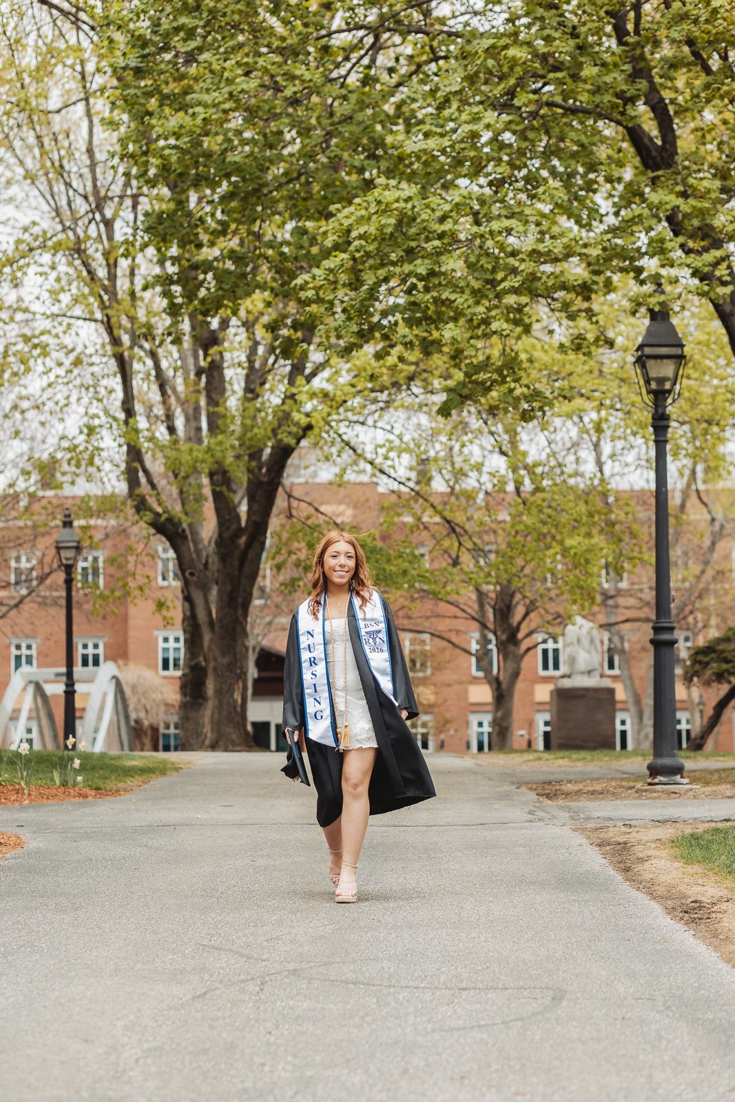 Young woman in graduation gown and sash walks on campus path surrounded by spring trees and brick buildings.