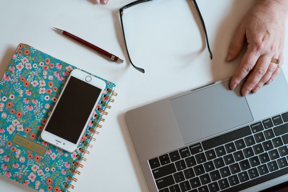 Overhead view of desk workspace with laptop, floral notebook, smartphone and glasses.