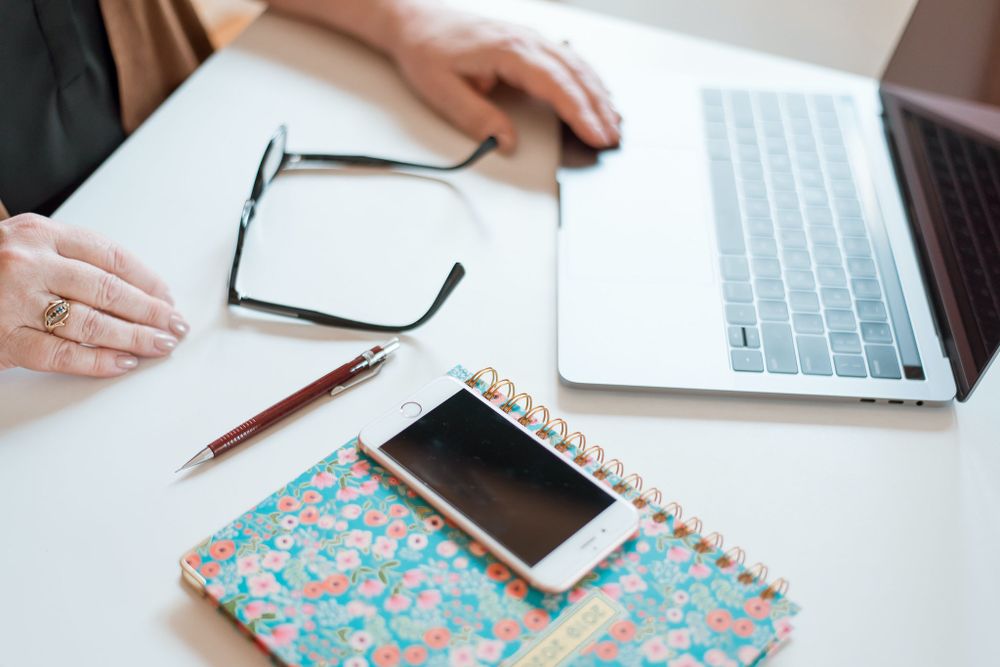 Close-up of hands at white desk with laptop, glasses, floral notebook and smartphone.