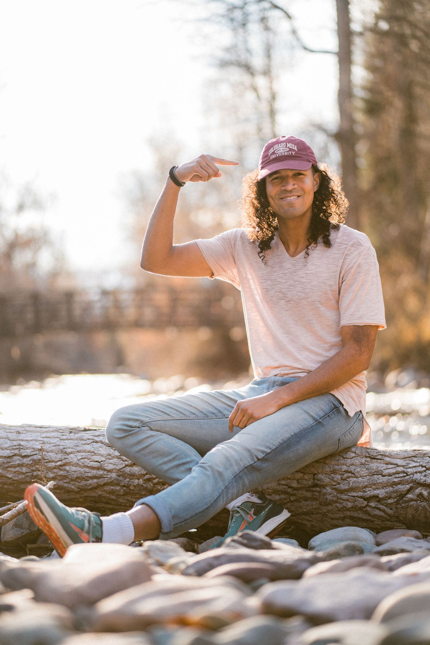 Young man with curly hair sitting on a log outdoors, wearing a pink tee, jeans, and a maroon cap, pointing at his hat.